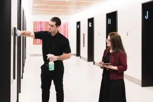 A man is cleaning a wall with spray while a woman observes, holding a tablet in a bright hallway.