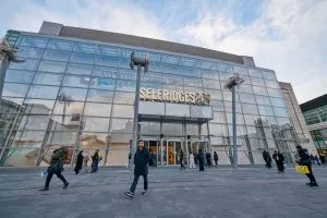 People walk past a glass-fronted store, reflecting the sky.