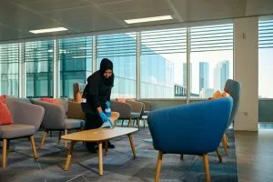 A person is cleaning a modern office lounge, wiping a table with a cloth, surrounded by colourful chairs and large windows.