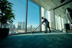A person is cleaning a large office window with a vacuum, overlooking a cityscape under a clear blue sky.