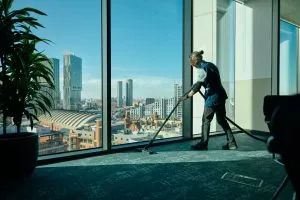 A person is hoovering a carpet in a high-rise office with a cityscape view through large windows, sunlight streaming in.